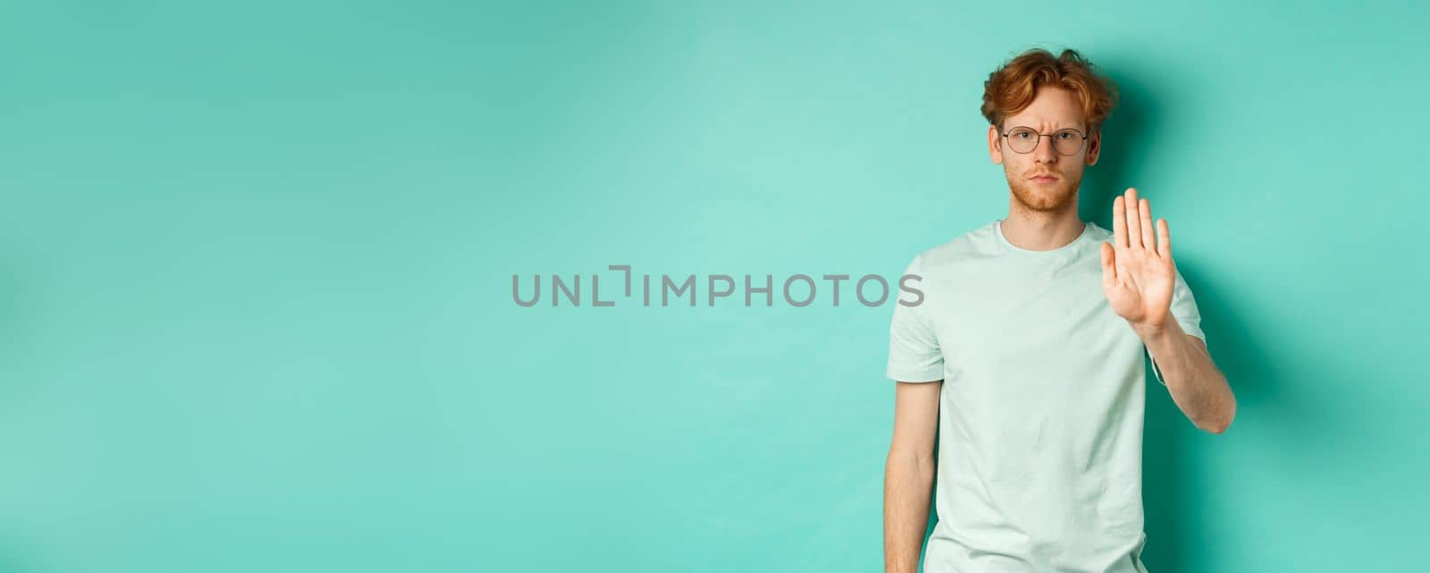 Angry and serious young man with red hair, wearing glasses, showing stop gesture, telling no, disapprove and prohibit something bad, standing over turquoise background by Benzoix