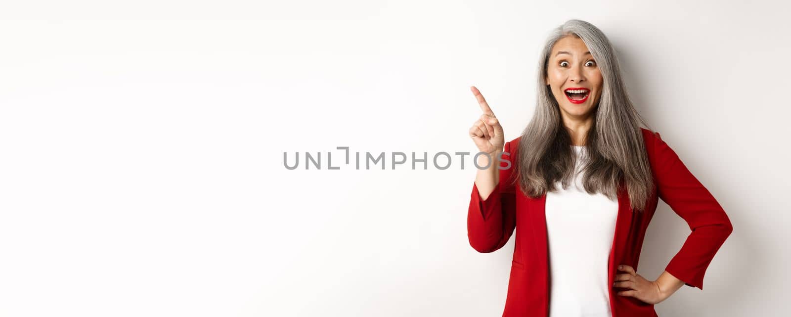 Business concept. Cheerful asian businesswoman with grey hair, wearing red blazer and makeup, pointing upper left corner and smiling amazed, white background.