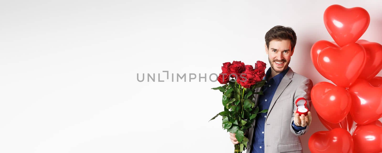 Handsome boyfriend in suit making a wedding proposal, showing engagement ring and say marry me, holding red roses, standing near Valentines day balloons, white background.