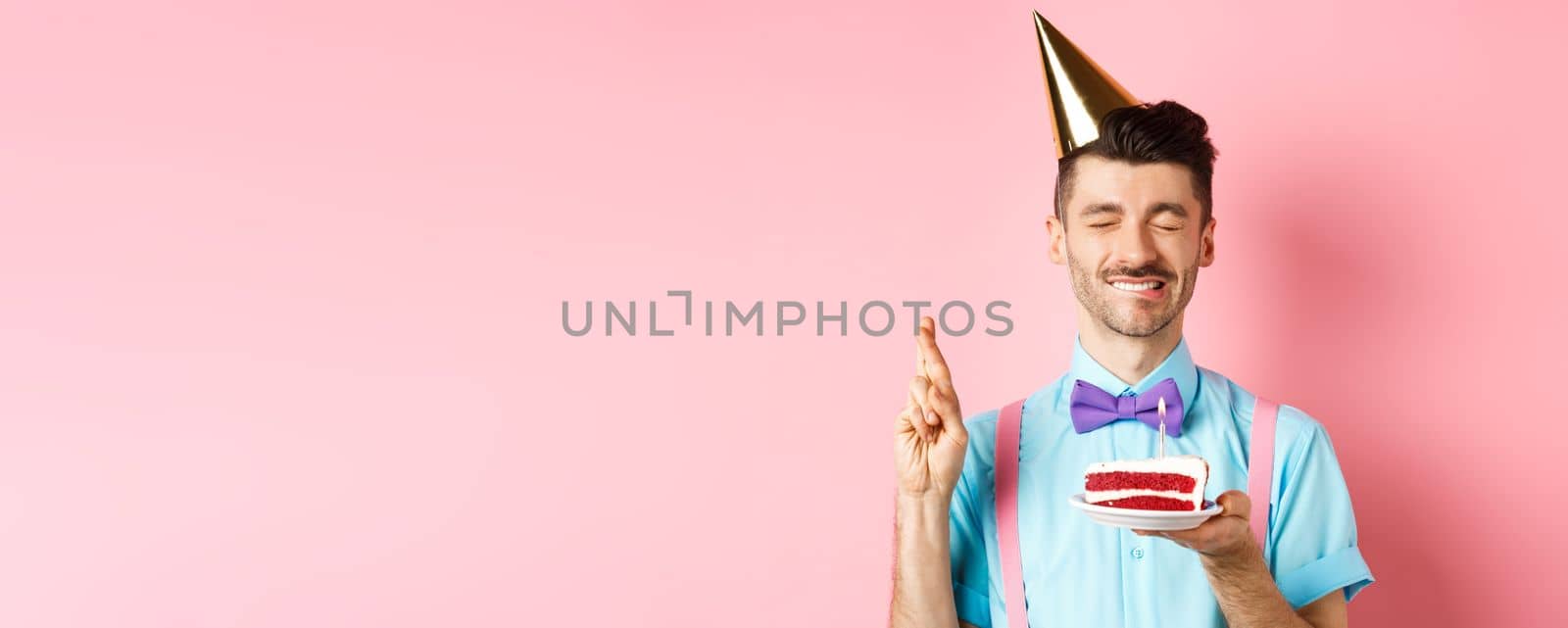 Holidays and celebration concept. Hopeful birthday guy in party hat making wish, holding cake with candle and cross fingers for dream come true, pink background.