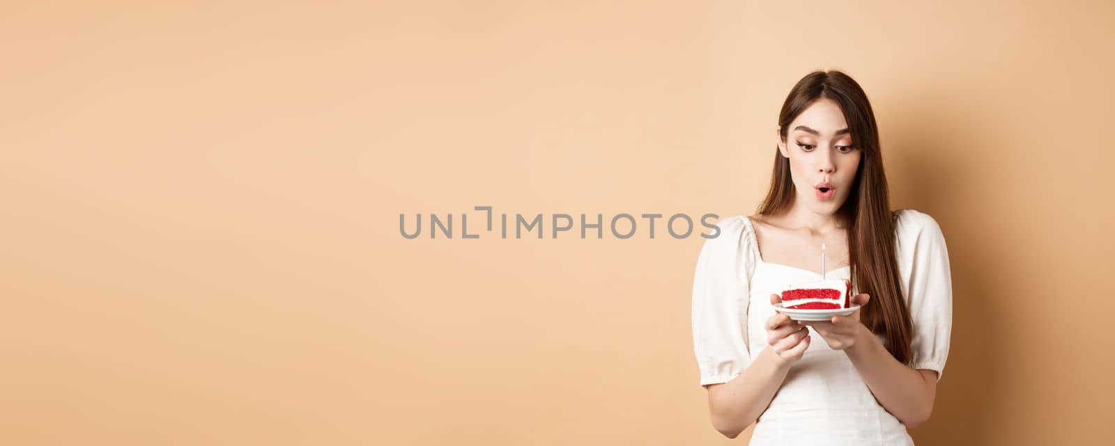 Beauty birthday girl blowing candle on b-day cake, making wish with excited face, standing on beige background and celebrating by Benzoix