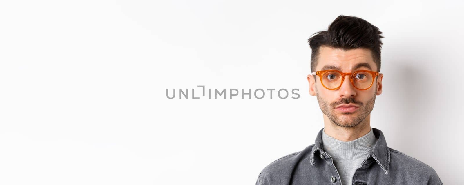 Attractive brunette man with moustache, wearing stylish glasses and look serious at camera, no emotion pokerface, standing against white background.
