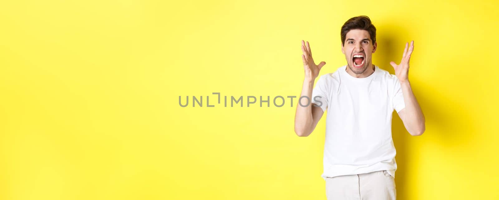 Angry man yelling and shaking hands, grimacing with hatred, standing hateful against yellow background by Benzoix