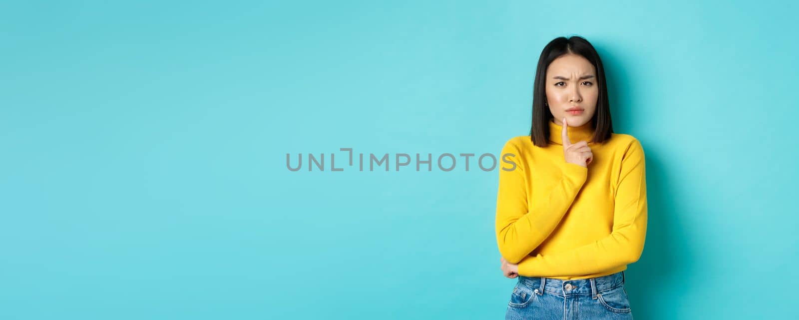 Image of pensive asian woman touching lip and frowning, thinking about something, trying to understand, standing over blue background.