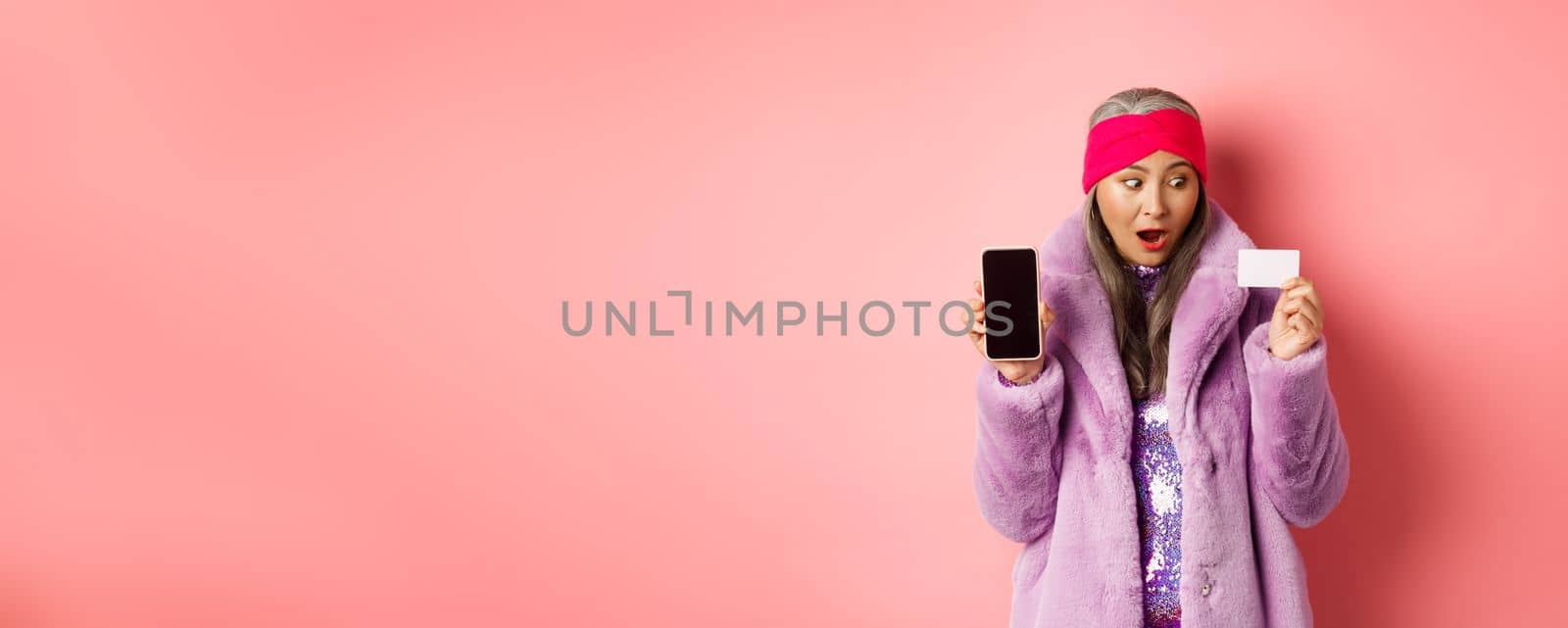 Special promotion. Excited asian senior woman showing blank smartphone screen and plastic credit card, checking out online offer, pink background.