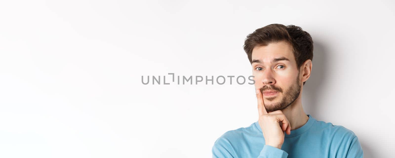 Image of thoughtful young man with beard, making choice, touching lip and looking pensive at camera, deciding something, white background.
