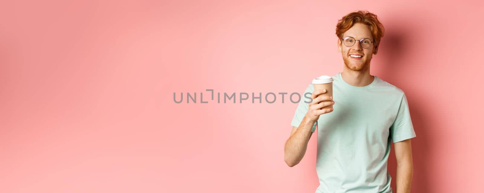 Happy redhead man in glasses and t-shirt drinking coffee and smiling, enjoying lunch break, holding takeaway cup, standing over pink background.