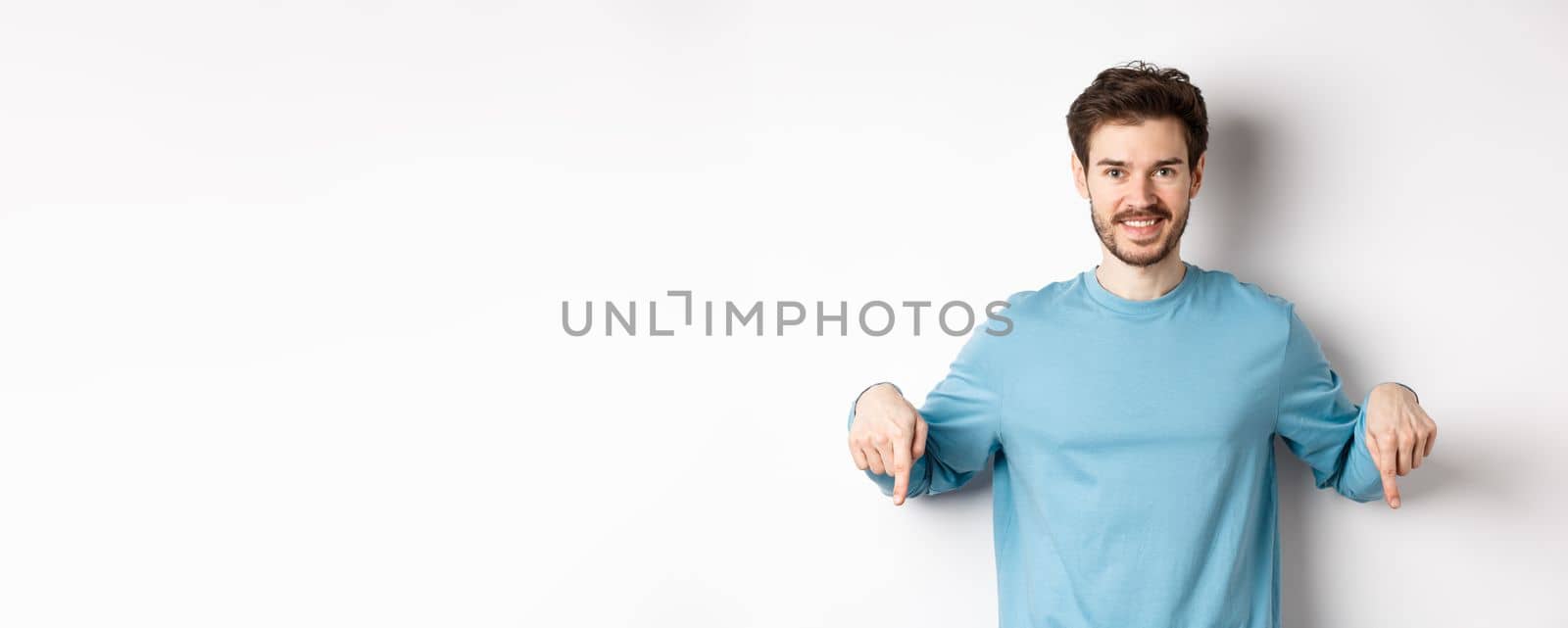 Smiling confident man pointing fingers down, showing promo banner or logo on white background, standing in casual blue sweatshirt.