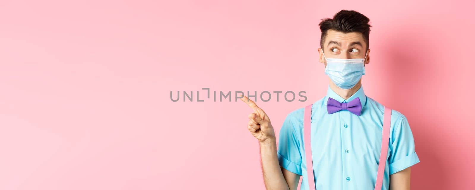 Covid-19, pandemic and health concept. Funny man in bow-tie and medical mask pointing finger left, looking aside curious, showing logo, standing on pink background.