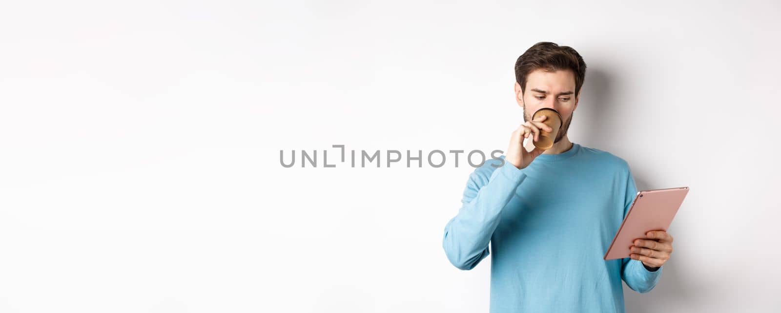 Handsome caucasian man drinking coffee and reading digital tablet screen, standing in blue sweatshirt over white background.