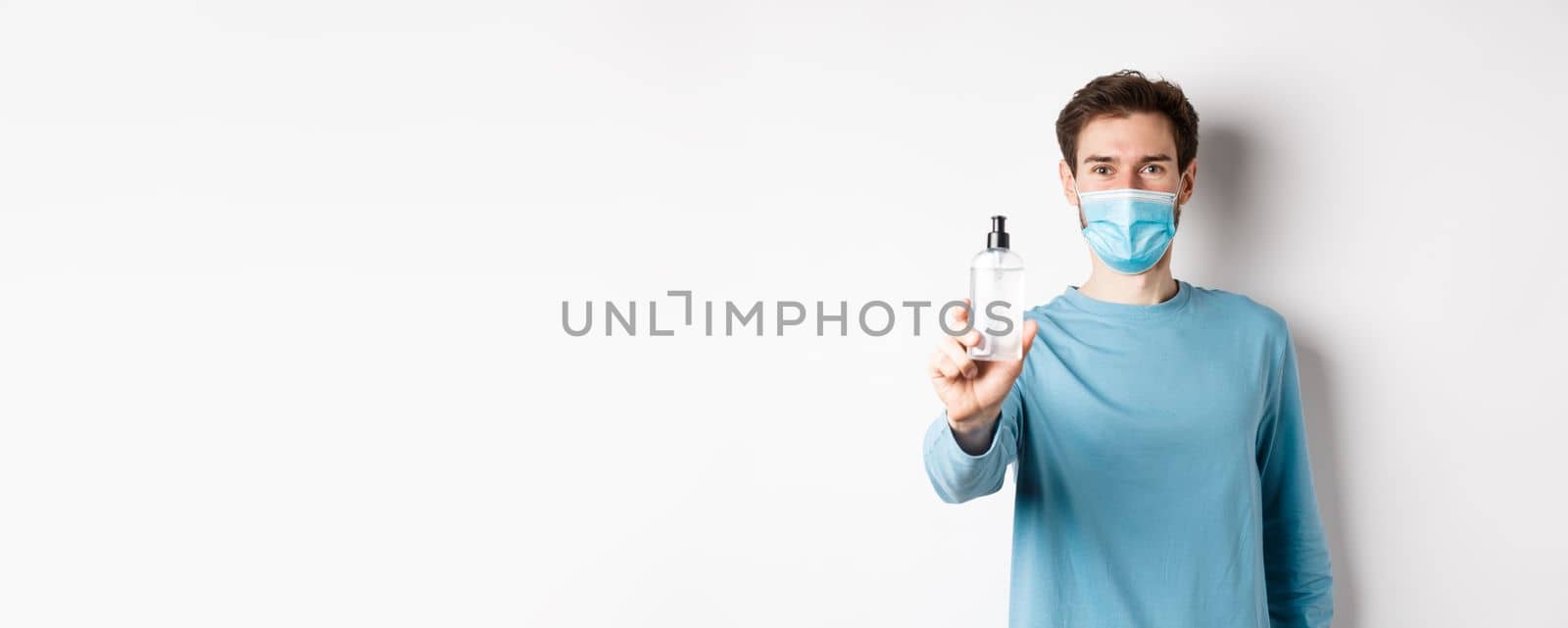 Covid-19, health and quarantine concept. Young man in medical mask stretch out hand and showing sanitizer, recommending antiseptic, standing over white background.