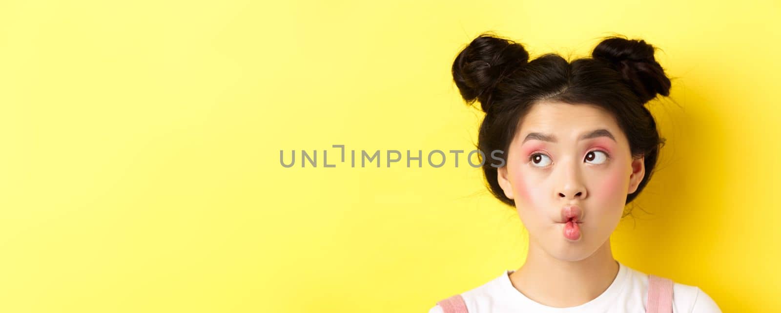 Close-up of teen asian girl pucker lips and looking funny at camera, standing with glamour makeup and stylish hairstyle, yellow background.