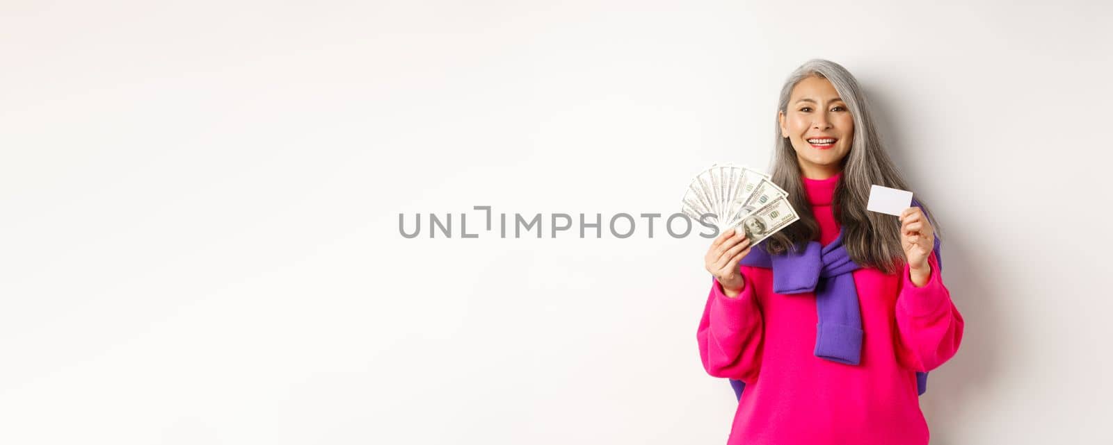 Shopping concept. Stylish asian senior woman with grey hair, showing money dollars and plastic credit card, standing over white background.
