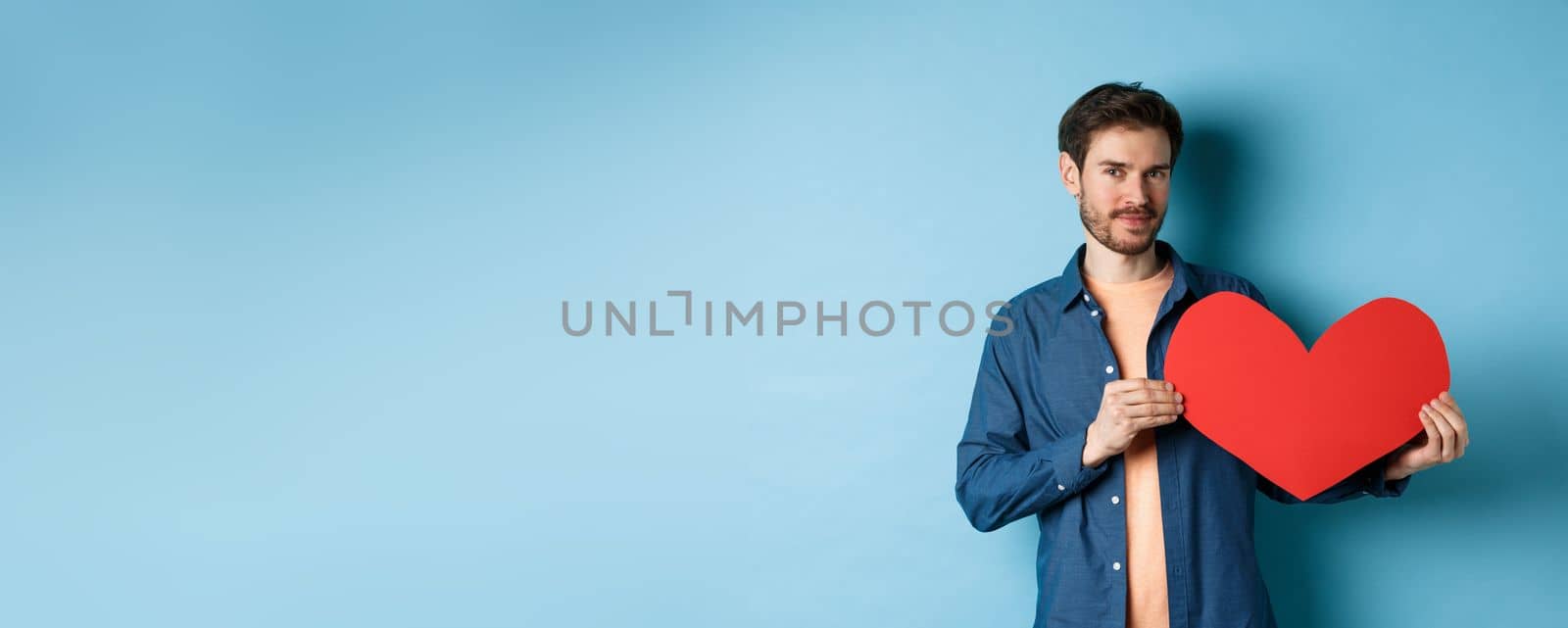 Handsome bearded guy showing valentines day cutout and smiling, standing in casual clothes over blue background. Copy space