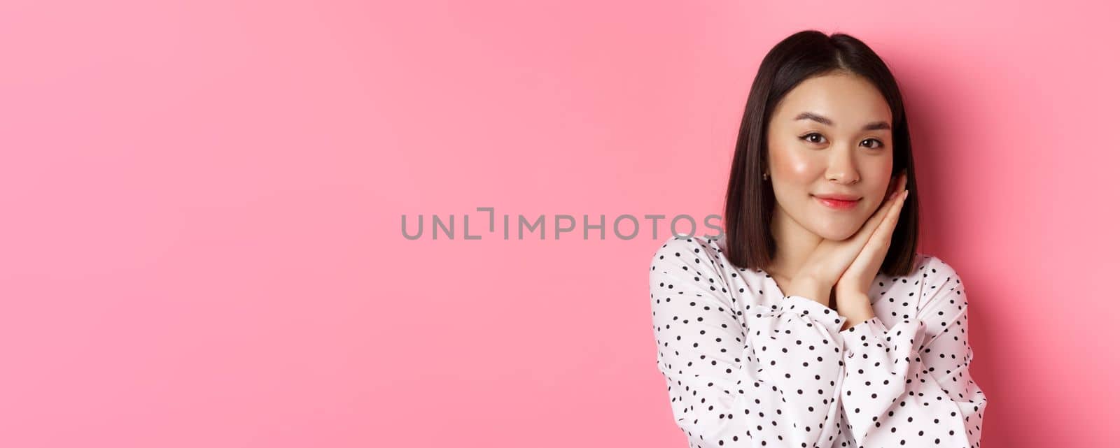 Beauty and lifestyle concept. Close-up of pretty korean girl leaning on palms, looking at camera with admiration, standing over pink background.