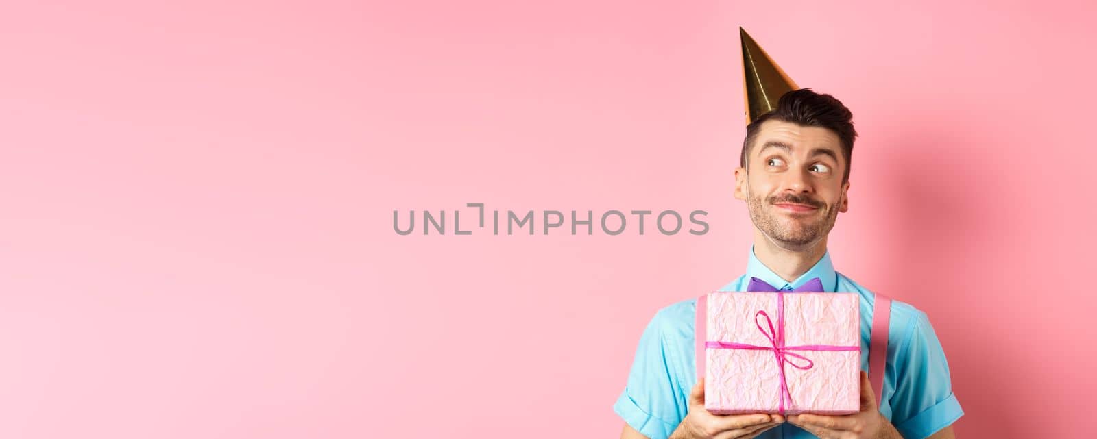 Holidays and celebration concept. Silly guy with moustache and bow-tie, wearing party hat, receive birthday gift and looking dreamy aside, standing over pink background.