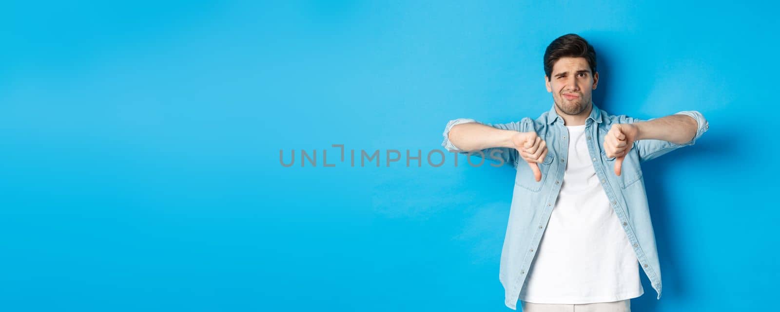 Skeptical and disappointed 25 years old man in casual outfit dislike something bad, showing thumbs-down and grimacing displeased, standing against blue background by Benzoix