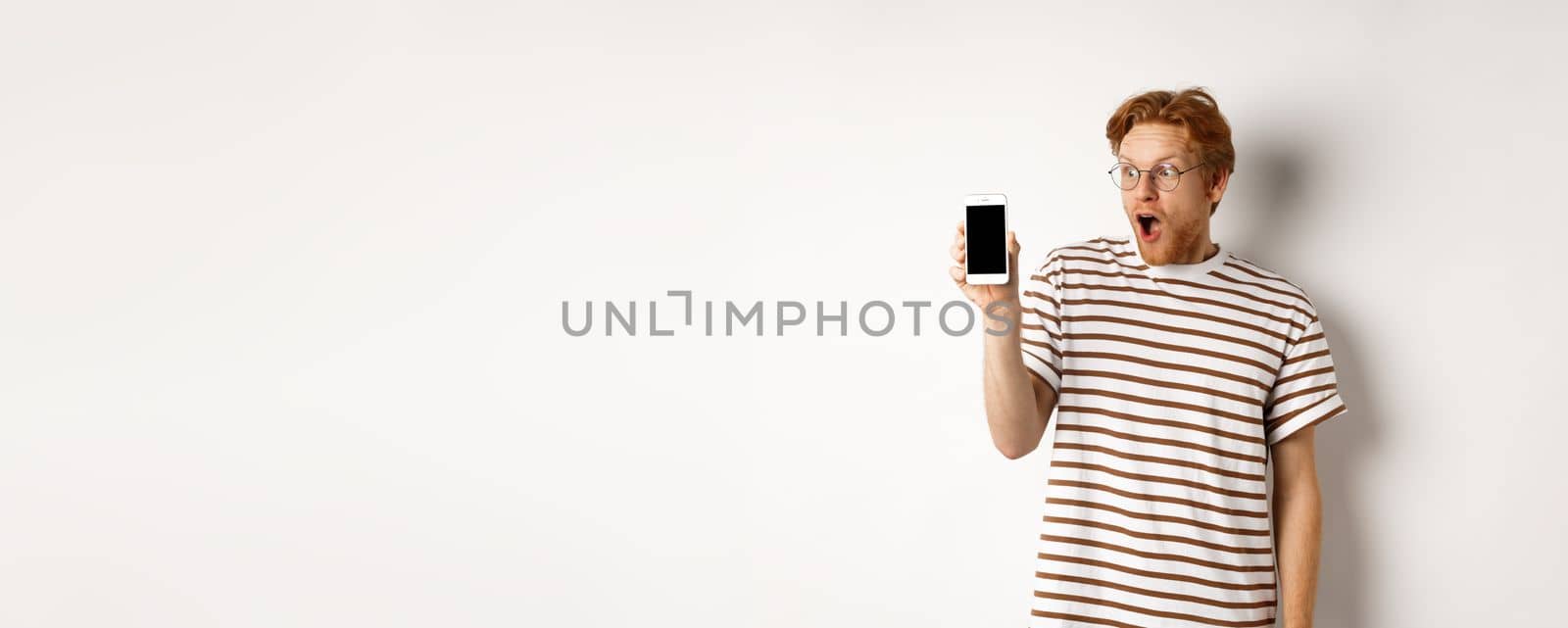 Technology and e-commerce concept. Surprised and shocked redhead guy checking out online promotion, showing blank smartphone screen and drop jaw, white background.