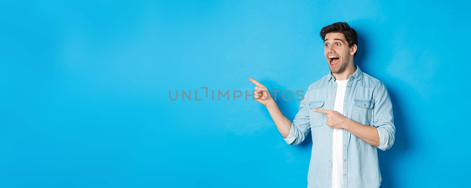 Portrait of handsome man looking and pointing fingers left with excitement, standing against blue background.