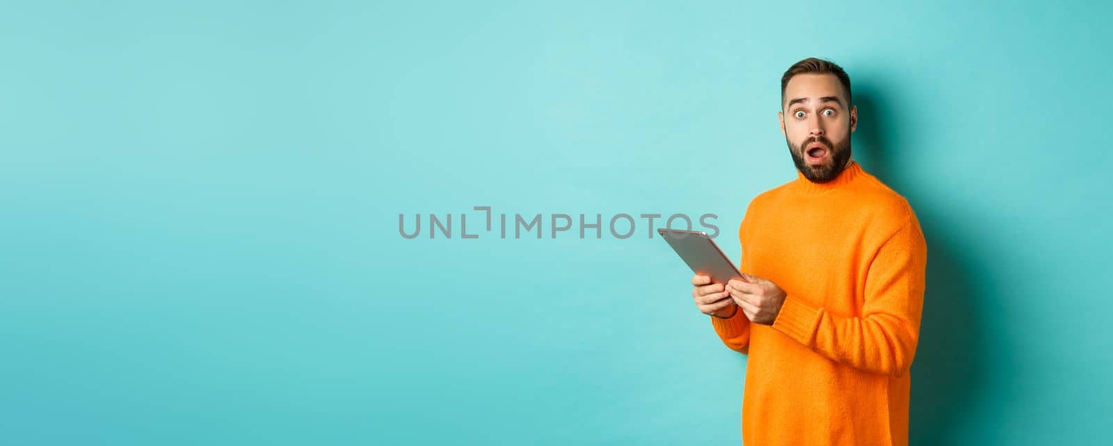 Image of male model in orange sweater using digital tablet, looking surprised, standing over light blue background.