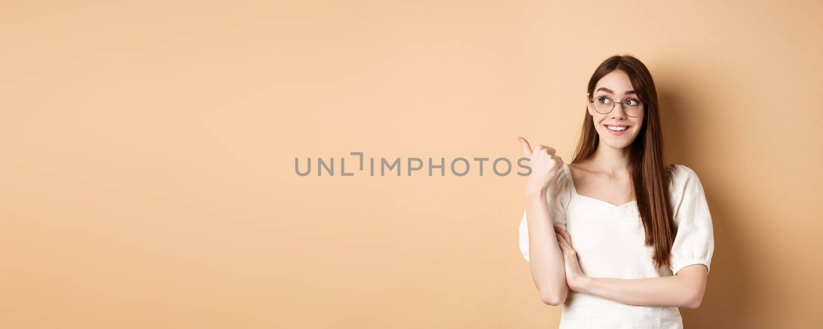 Happy woman in glasses smiling and pointing left, showing logo on empty space, standing on beige background by Benzoix