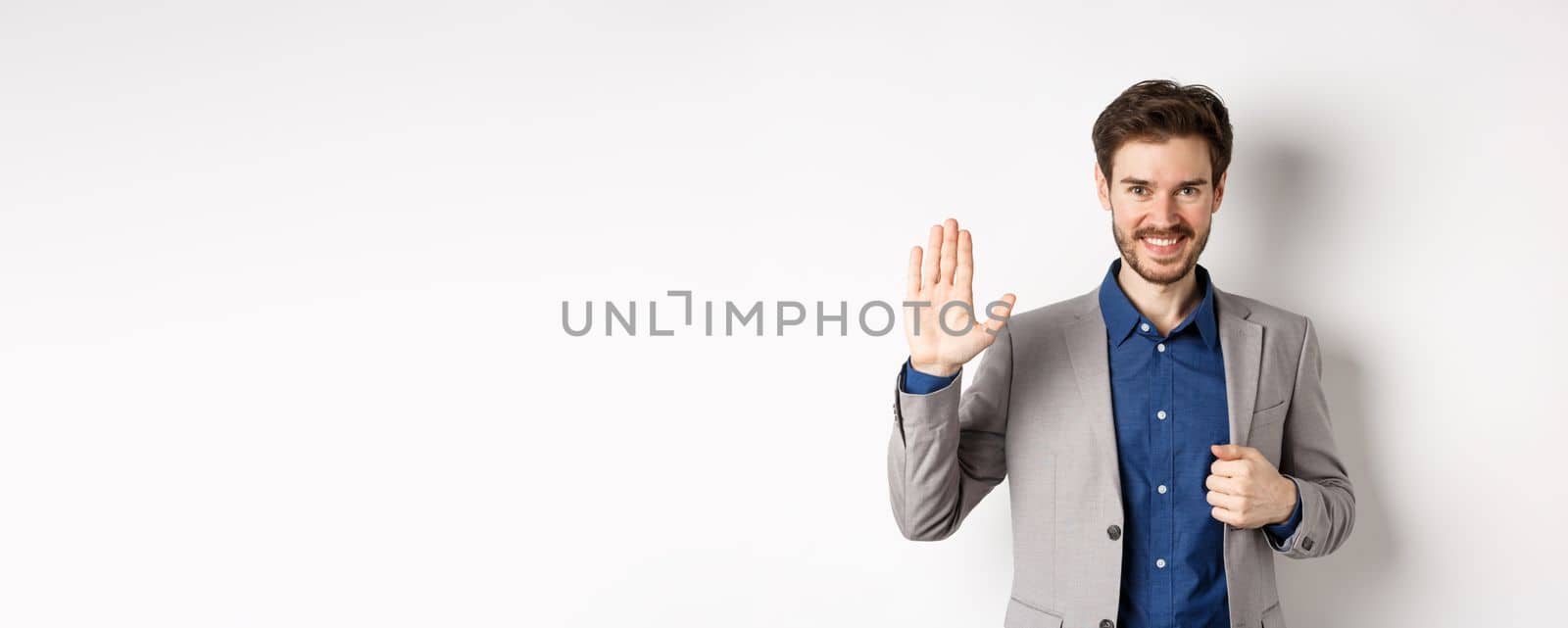 Smiling handsome businessman in suit waving hand, greeting friend with friendly face, say hellp, standing on white background.