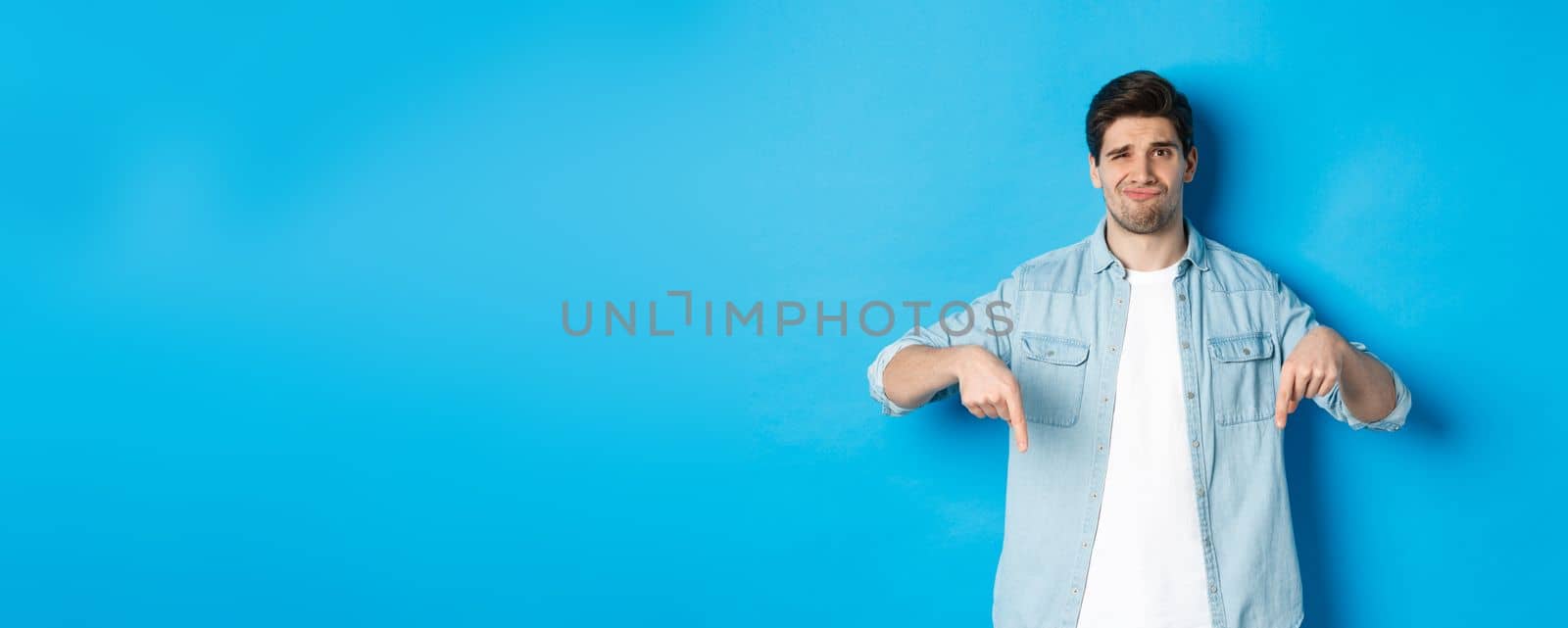 Disappointed guy in casual outfit, pointing fingers down with doubtful grimace, standing against blue background.