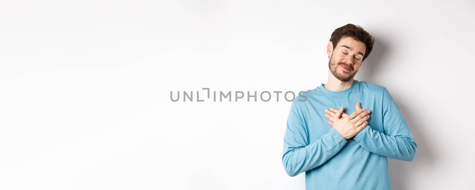 Handsome young man feeling romantic, holding hands on heart and thinking about good pleasant memories, standing over white background by Benzoix