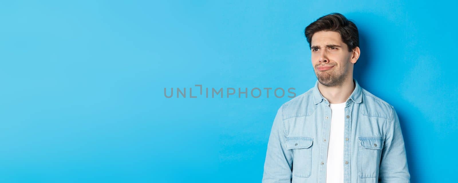 Close-up of upset and disappointed man grimacing, looking dissatisfied left, cringe from bad promo banner, standing over blue background.