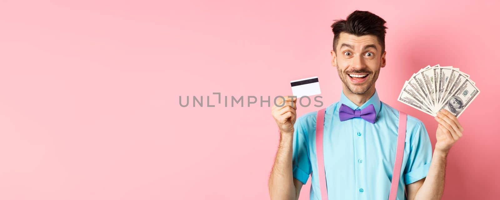 Cheerful caucasian man with moustache and bow-tie showing plastic credit card with money in dollars, smiling at camera, standing over pink background.