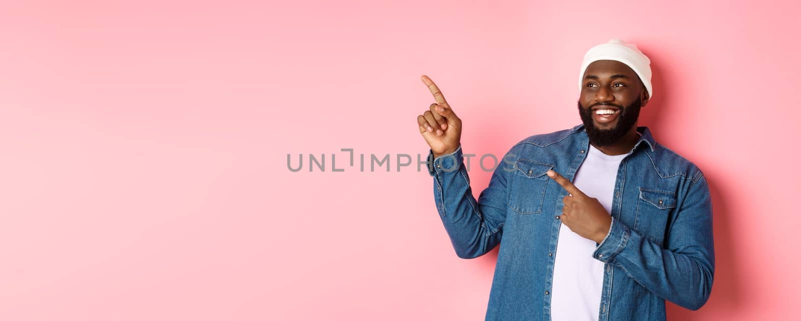 Amazed Black guy showing awesome promo offer, smiling and pointing fingers at upper left corner banner, pink background by Benzoix