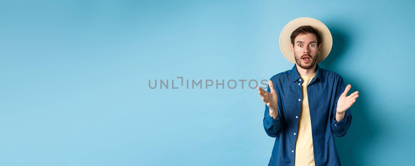 Shocked male tourist on vacation raising hands up and looking at camera startled, react to big news with disbelief, standing on blue background in straw summer hat and shirt by Benzoix