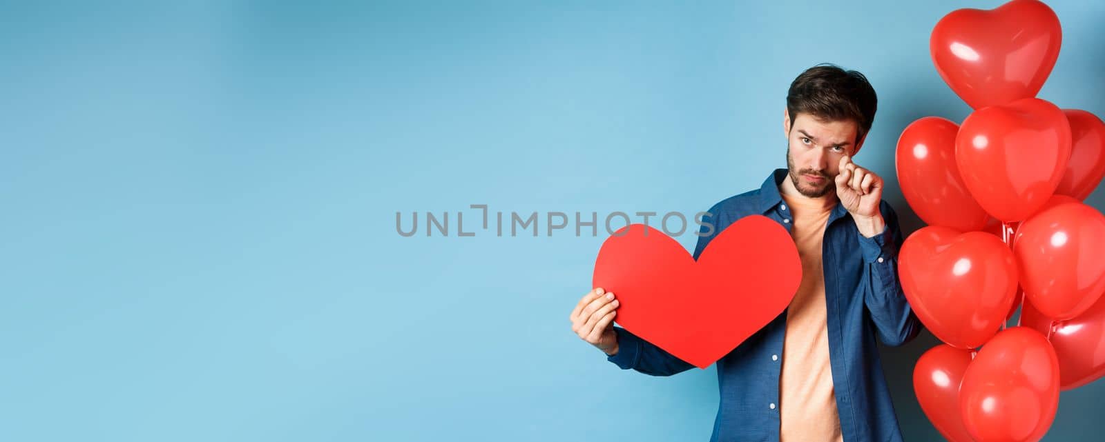 Sad and heartbroken man crying, wiping tears, standing with red heart and balloons, breakup on Valentines day, blue background.