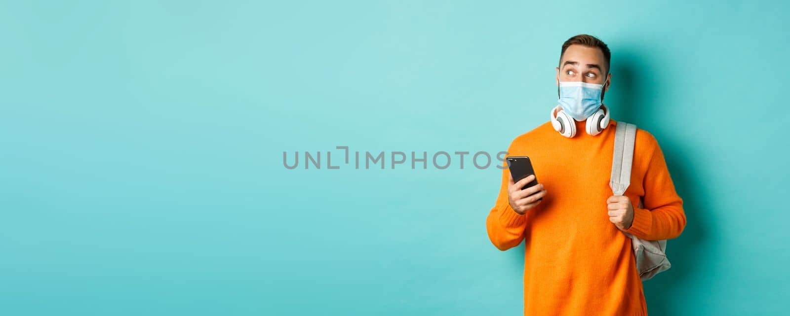 Young man in face mask using mobile phone, holding backpack, staring left amazed, standing against light blue background by Benzoix