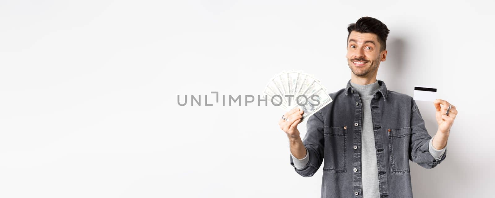 Happy guy showing plastic credit card and dollar bills, giving choice, standing on white background by Benzoix