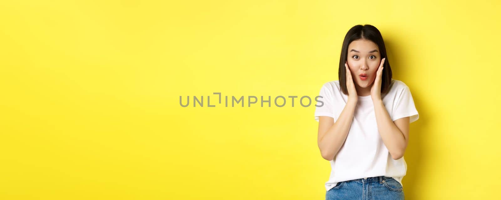 Beauty and fashion concept. Asian girl saying wow with amazement, checking out awesome promo deal, standing over yellow background.