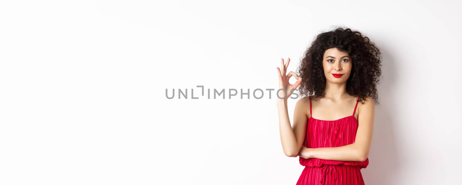 Beautiful assertive woman with curly hairstyle and makeup, wearing red dress, showing okay sign and smiling in approval, say yes, standing on white background.