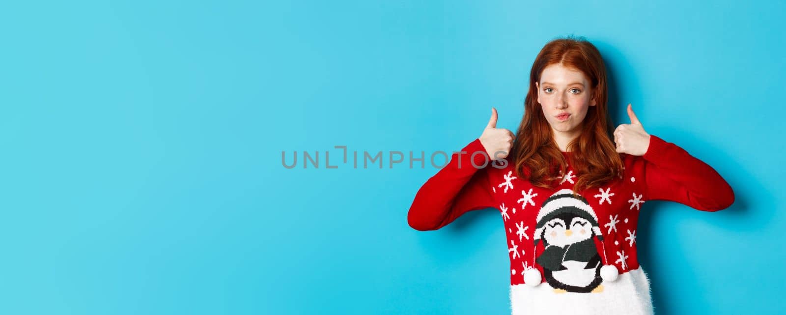 Winter holidays and Christmas Eve concept. Impressed redhead girl in xmas sweater, nod in approval and showing thumb up, praise good product, standing over blue background.