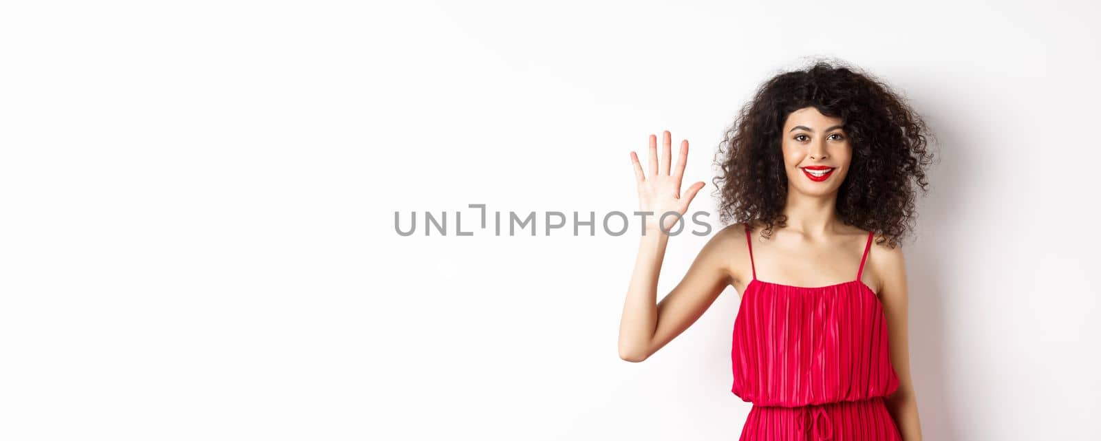 Cheerful young woman with makeup and red dress, showing five fingers and smiling, standing over white background. Copy space