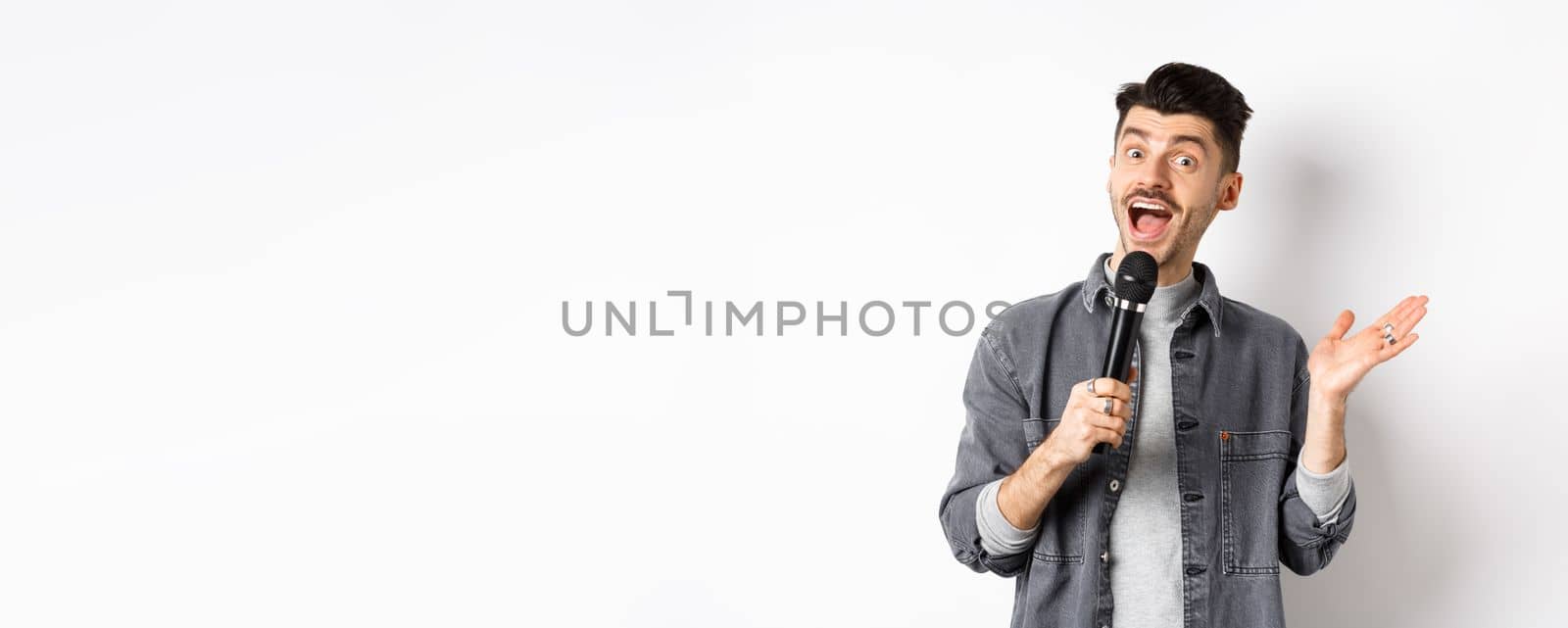 Excited stylish guy giving speech, talking in microphone, perform or sing karaoke, standing on white background.