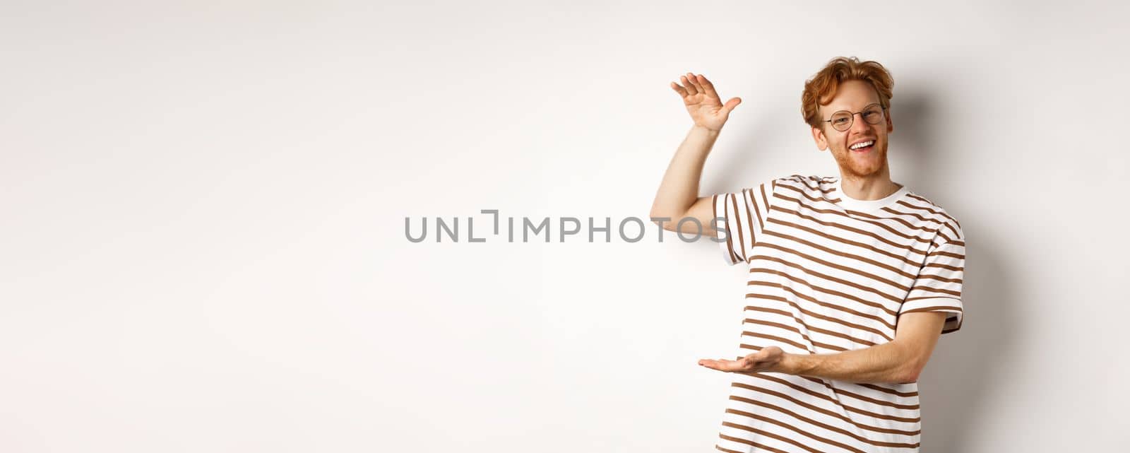 Happy and proud redhead guy with glasses showing something big, shaping large object with hands over white background, smiling at camera by Benzoix