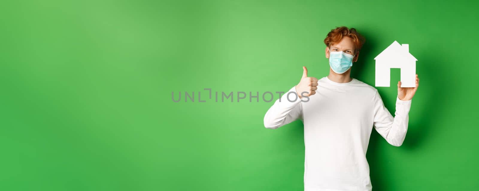 Real estate and covid-19 pandemic concept. Young redhead man in medical mask showing paper house cutout and showing thumb-up in approval, recommending agency by Benzoix