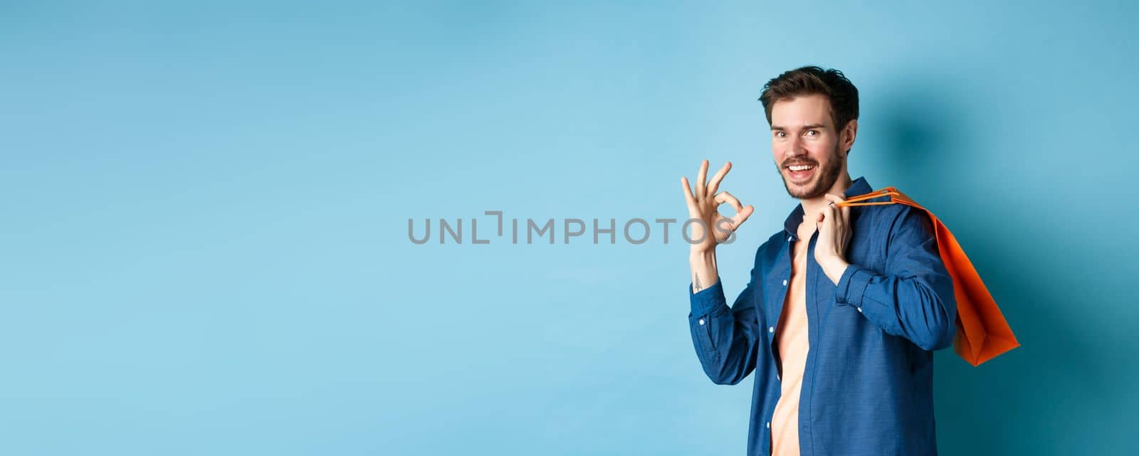Handsome young guy with shopping bag showing okay sign and smiling, praising good deal, standing on blue background.