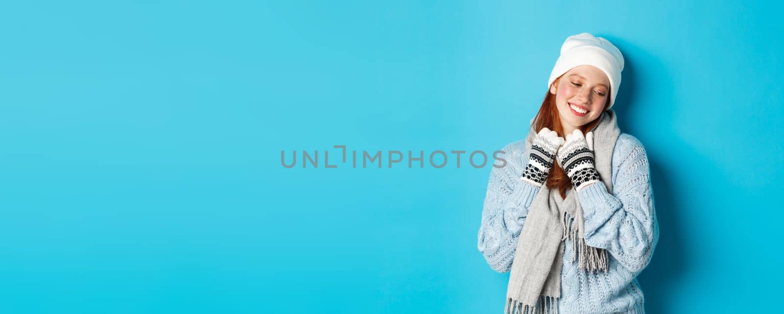 Winter and holidays concept. Cute redhead girl blushing and looking away dreamy, wearing warm beanie, sweater and scarf with gloves, standing over blue background by Benzoix
