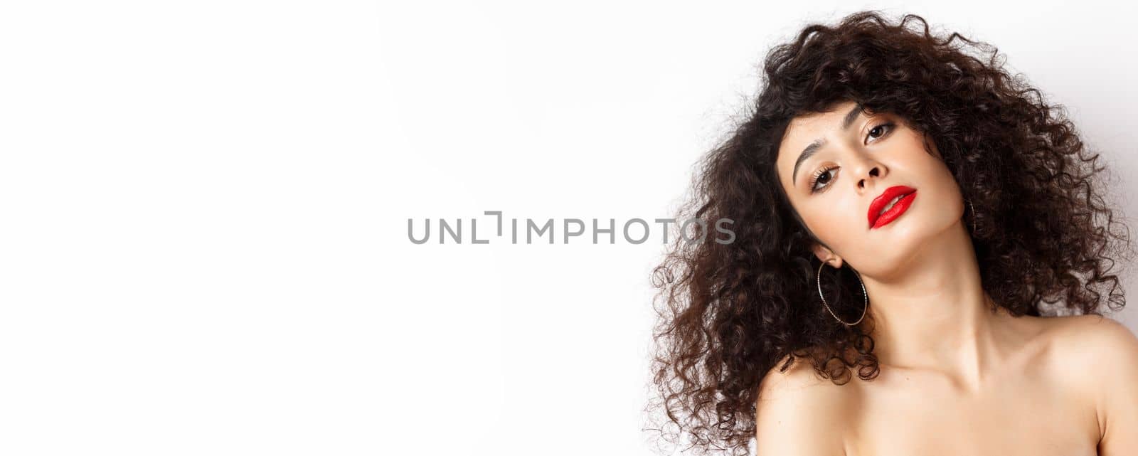 Close-up portrait of sensual and romantic woman with curly hair, red lips, looking seductive at camera, posing on white background.