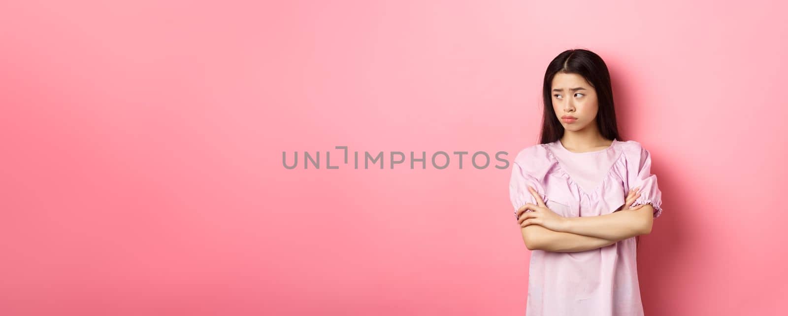 Sad and gloomy asian woman look away at logo, sulking from unfair situation, cross arms on chest disappointed, standing against pink background by Benzoix