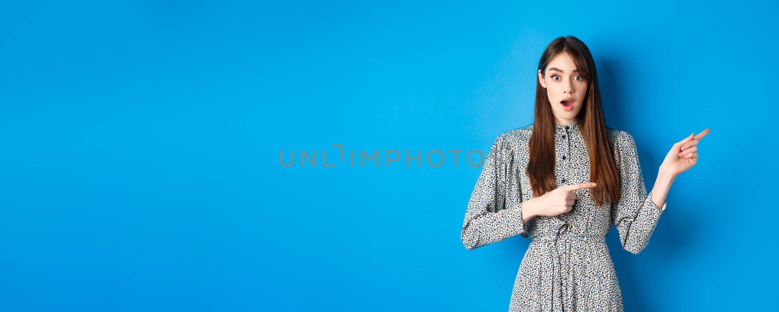 Wow look that way. Excited young woman with long hair and trendy dress, drop jaw and pointing right at logo, showing awesome news, blue background by Benzoix