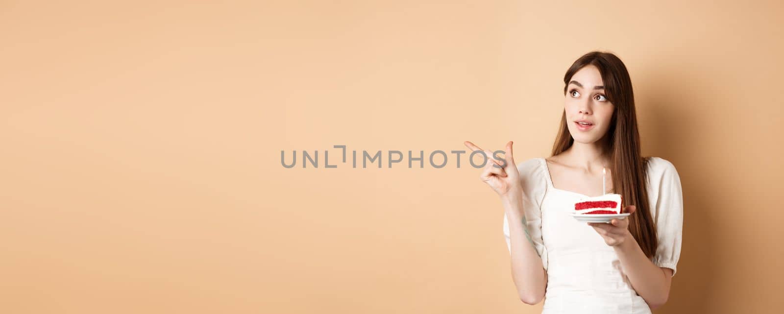 Thoughtful birthday girl holding cake with candle, thinking of wish and pointing left at logo, standing on beige background.