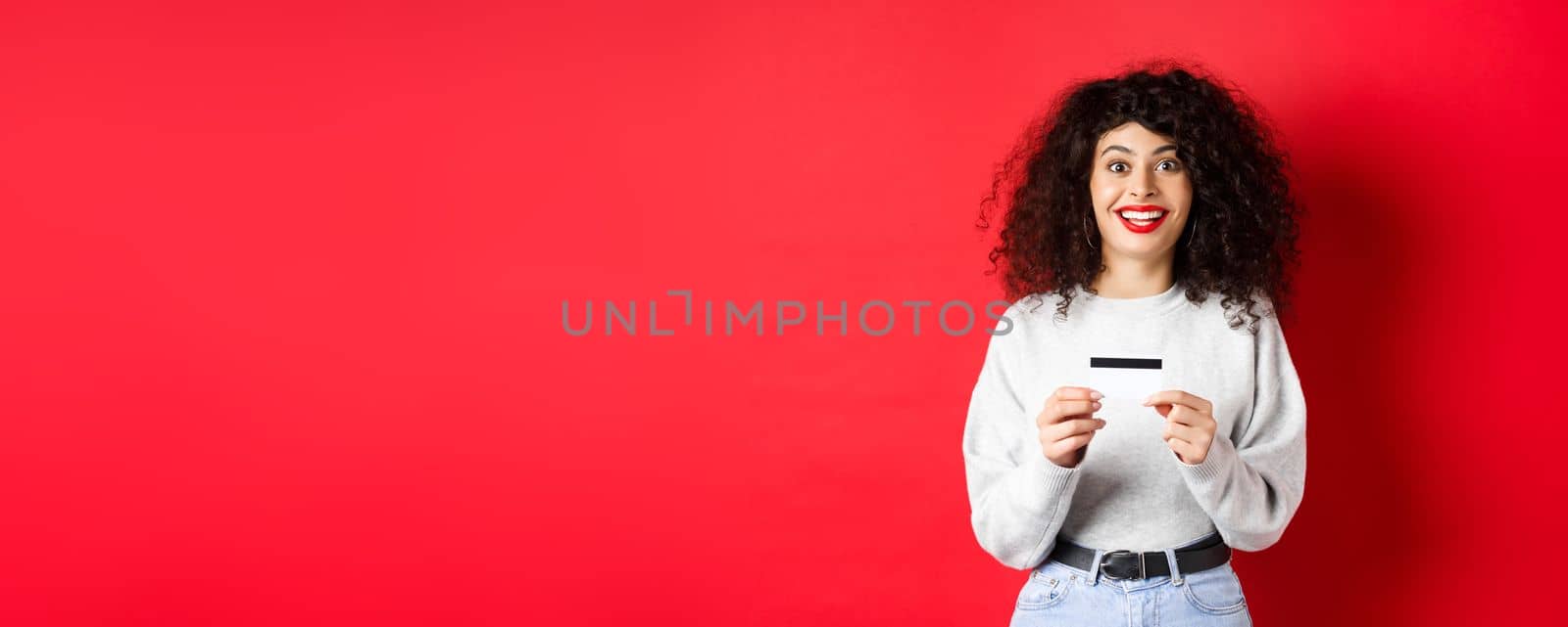 Excited smiling woman showing plastic credit card, standing in casual clothes on red background.