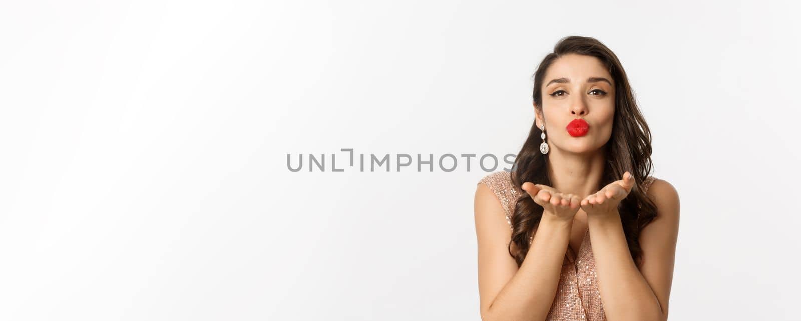 Concept of New Year celebration and winter holidays. Close-up of sensual young woman in dress, pucker lips and blowing air kiss at camera, white background.